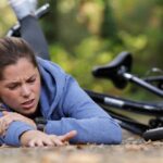 Frau liegt nach Fahrradsturz am Boden, Fahrrad im Hintergrund, Gesicht schmerzverzerrt